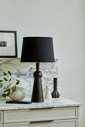 Black solid wood table lamp with gently tapered body and black linen shade in kitchen scene with candle holders and art objects