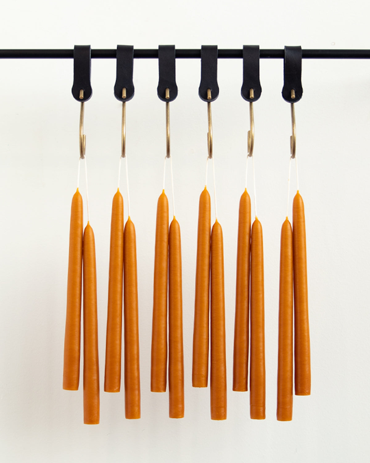 A dozen orange taper candles hanging in pairs from black leather hooks on a black wall pot rack.