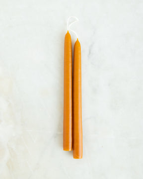A pair of hand dipped creamy orange taper candles on a lightly marbled white background.