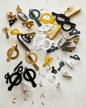 Hooks for wall storage in brass, black, white, mustard and smoke and multiple sizes scattered with brass hardware on a white textured background.