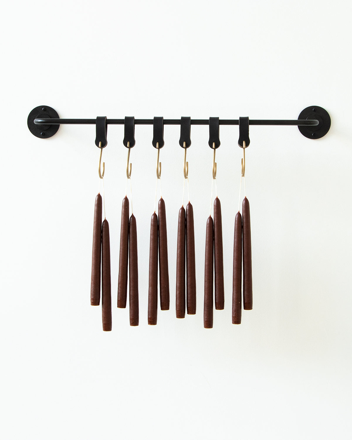 A dozen chestnut brown taper candles hanging in pairs from black leather hooks from a black wall pot rack.