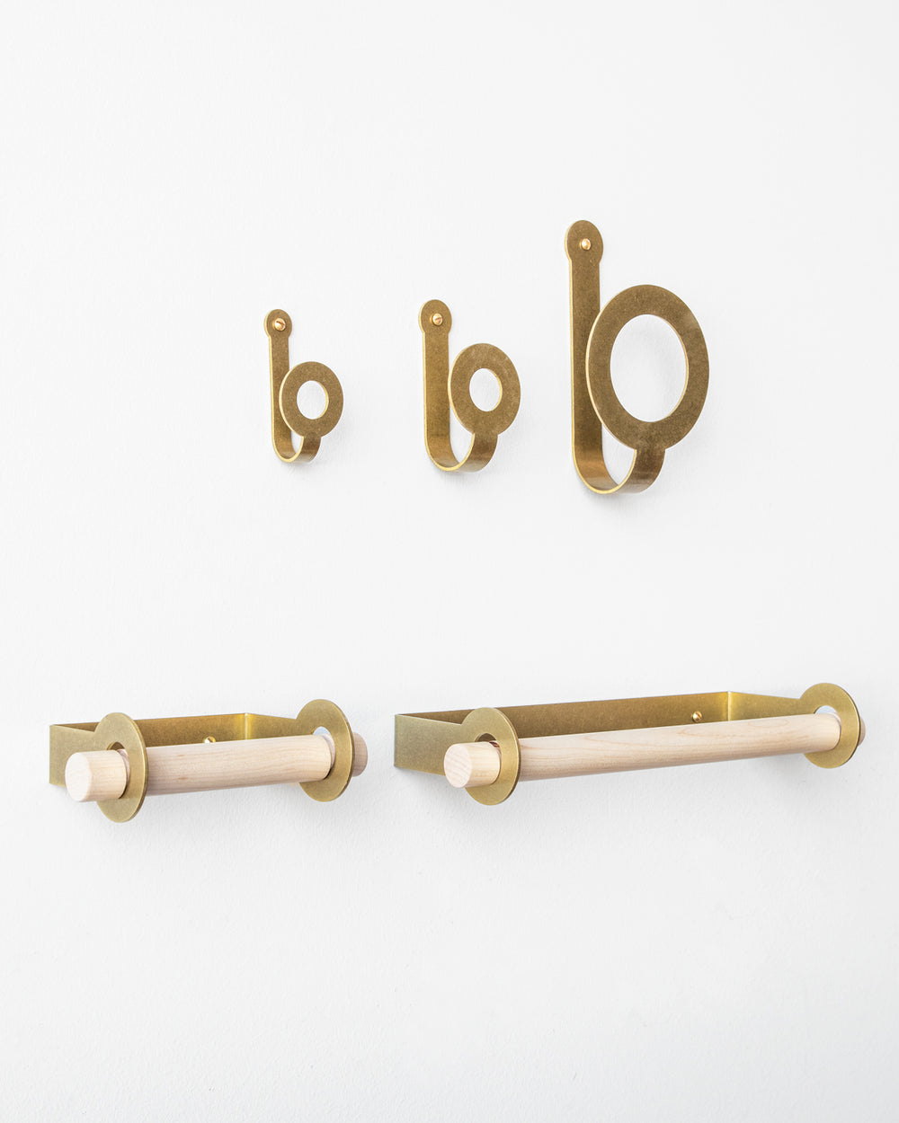 Three sizes of brass wall hooks above a brass toilet paper holder and brass paper towel holder, both empty. #finish_brass