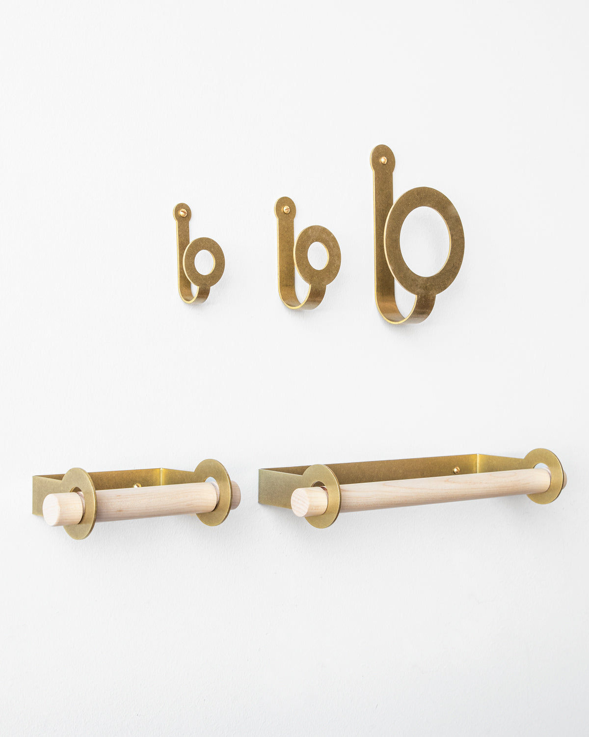 Three sizes of brass wall hooks above a brass toilet paper holder and brass paper towel holder, both empty.