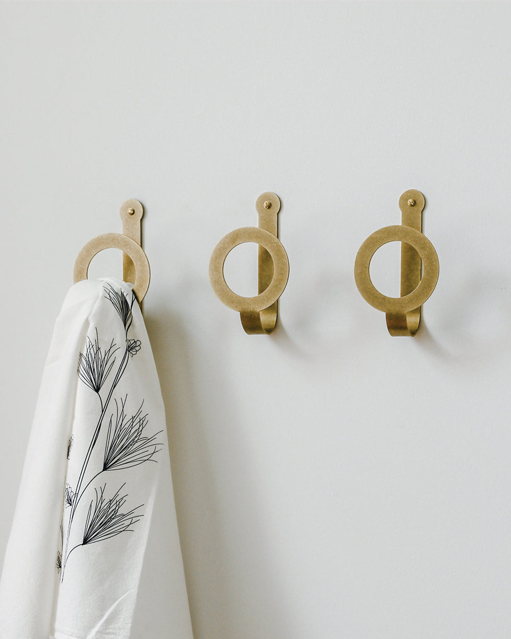 Three oversized brass wall hooks in a line. One holds a white towel patterned with black flowers. #finish_brass