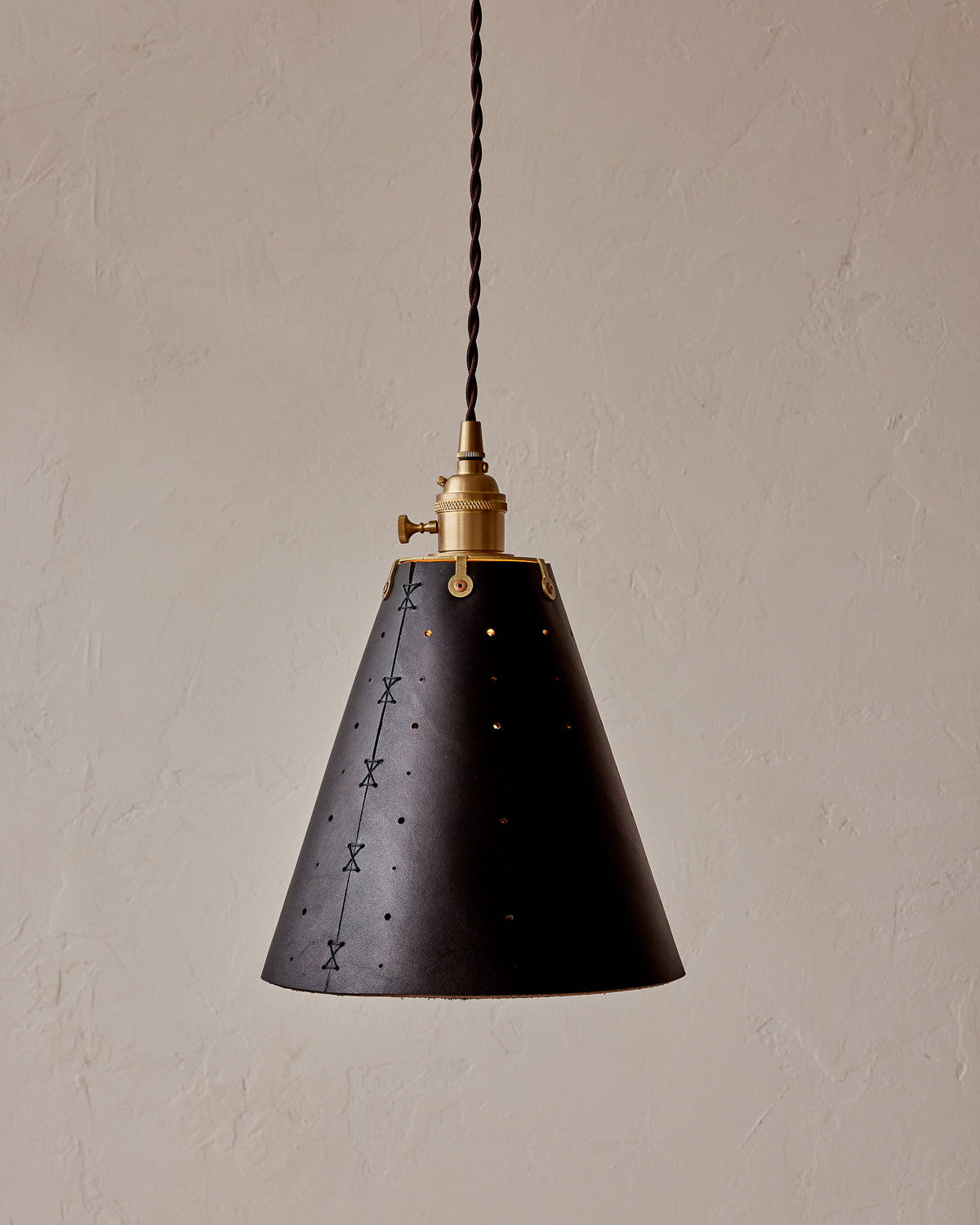 A black pendant light with a conical shade featuring patterned perforations, a brass fitter, and a twisted cloth cord. #color_black harness